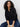 A woman with curly hair sits on a chair, calmly looking at the camera in Sanctuary Clothing's petal drop blouse in black and dark blue jeans, against a plain light background.