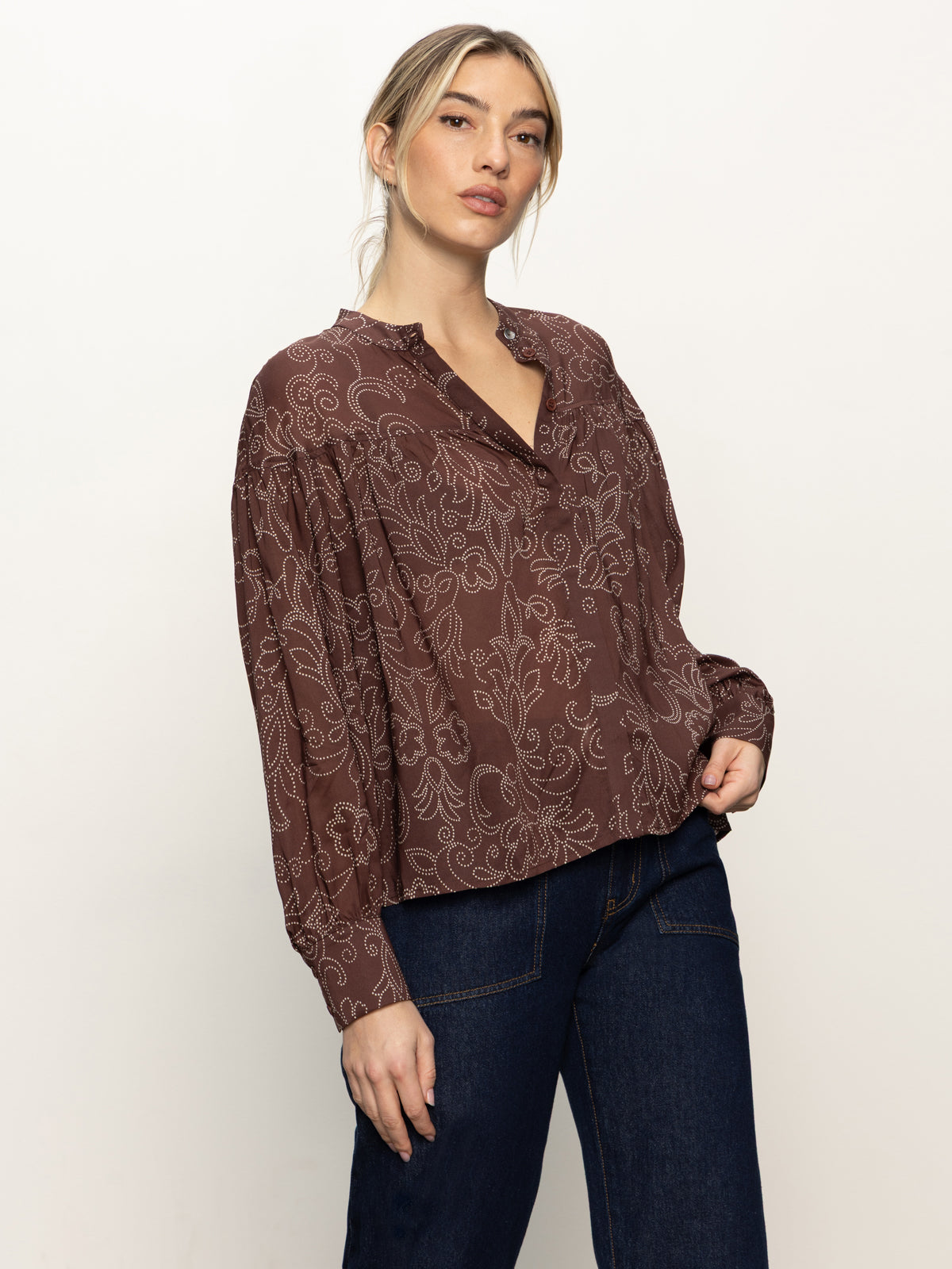 Someone wears the Sanctuary Clothing personal best blouse boheme—a loose, long-sleeve brown top with a white floral pattern—paired with dark blue jeans, standing against a plain light background.