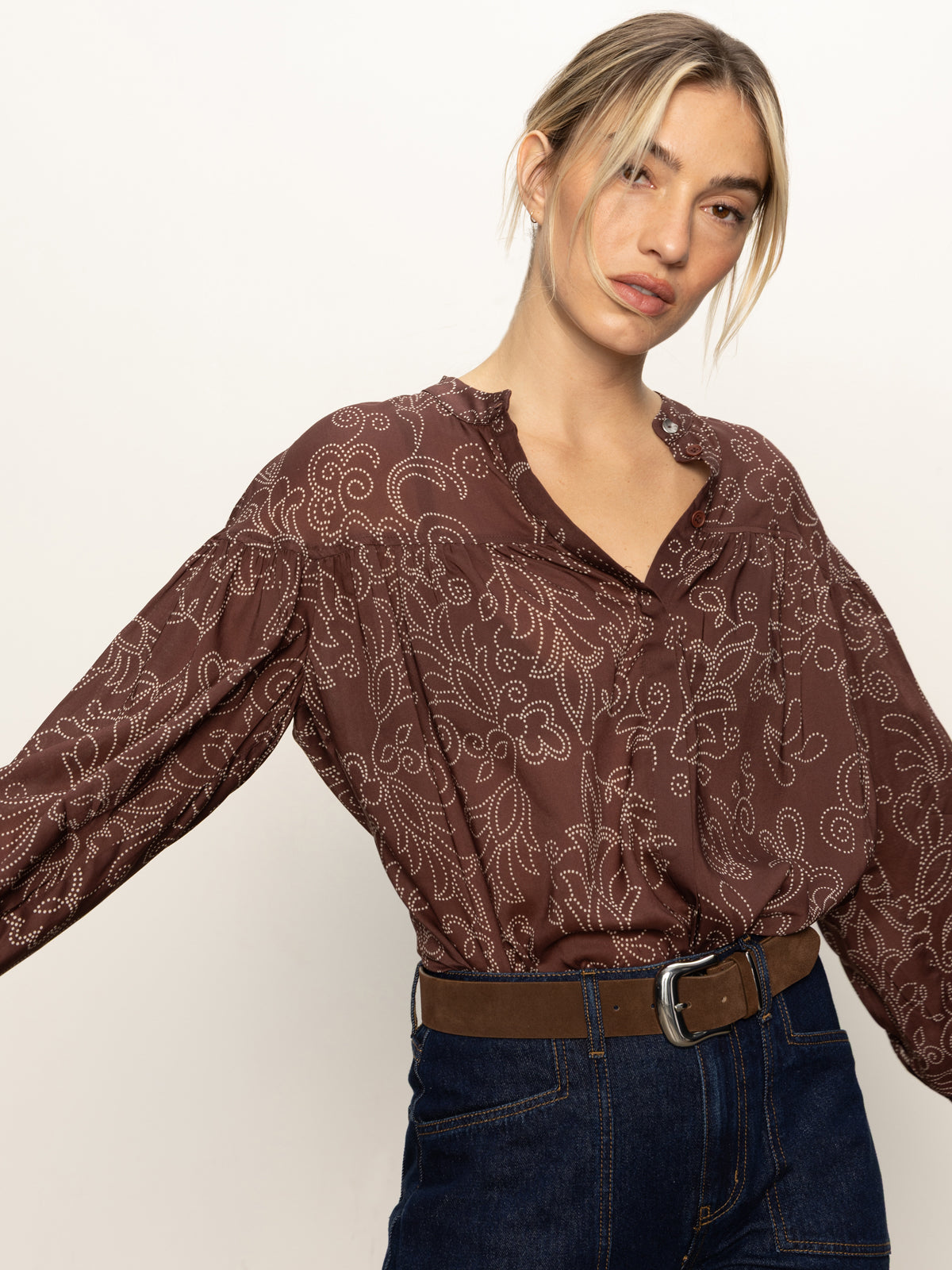A woman with blonde hair tied back wears the Sanctuary Clothing personal best blouse boheme, featuring a white floral pattern, paired with dark jeans and a brown belt. She looks toward the camera with a neutral expression.