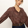 A woman with blonde hair tied back wears the Sanctuary Clothing personal best blouse boheme, featuring a white floral pattern, paired with dark jeans and a brown belt. She looks toward the camera with a neutral expression.