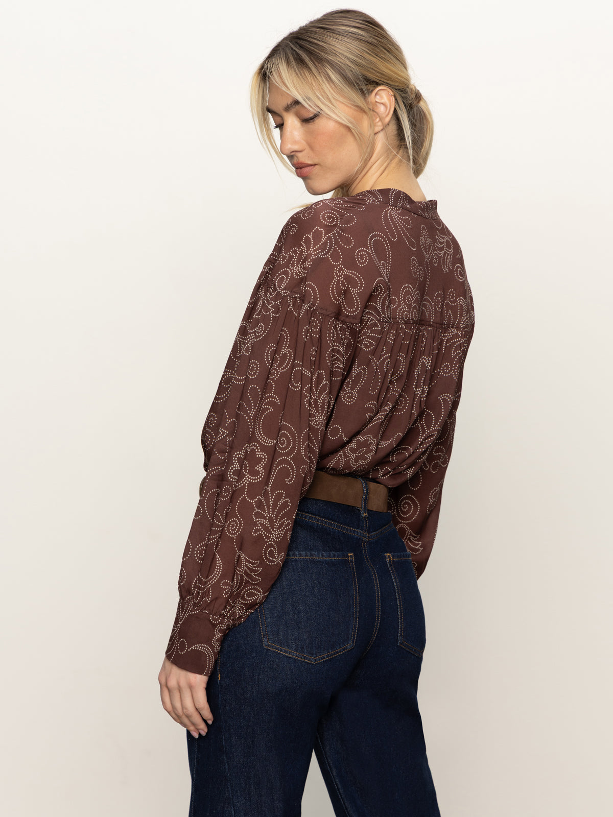 A woman with blonde hair in a low ponytail wears the Sanctuary Clothing personal best blouse boheme and dark denim jeans, standing sideways and looking down over her shoulder against a plain background.