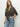 A woman with long blonde hair models the Sanctuary Clothing Wild Love Tie Top French Kitty—a leopard print blouse tied at the waist—paired with light blue high-waisted jeans, standing against a plain white background.