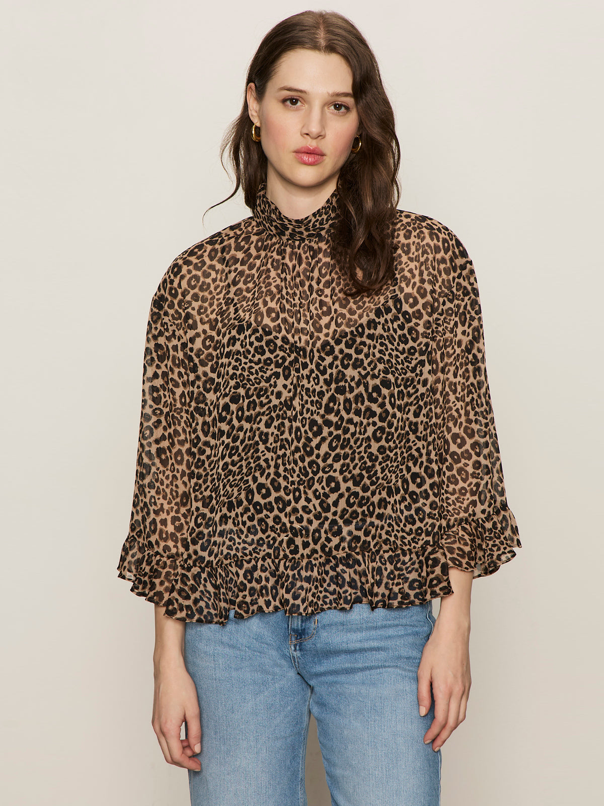 A woman with long brown hair wears the adore blouse french kitty by Sanctuary Clothing—a sheer leopard print top with ruffled cuffs—paired with blue jeans, standing against a plain light background.