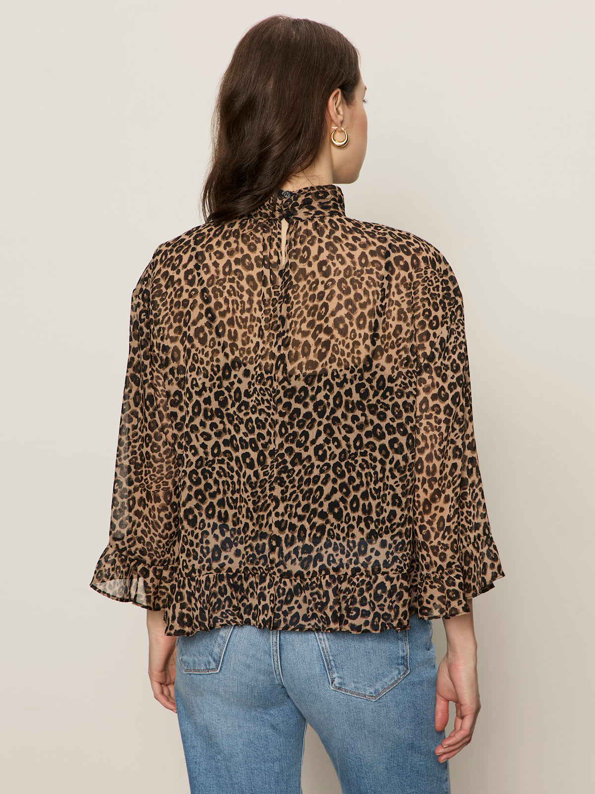 A woman with long brown hair wears gold hoop earrings, light blue jeans, and Sanctuary Clothing's the adore blouse french kitty—a sheer, high-neck leopard print top with a keyhole back—standing against a plain light background.