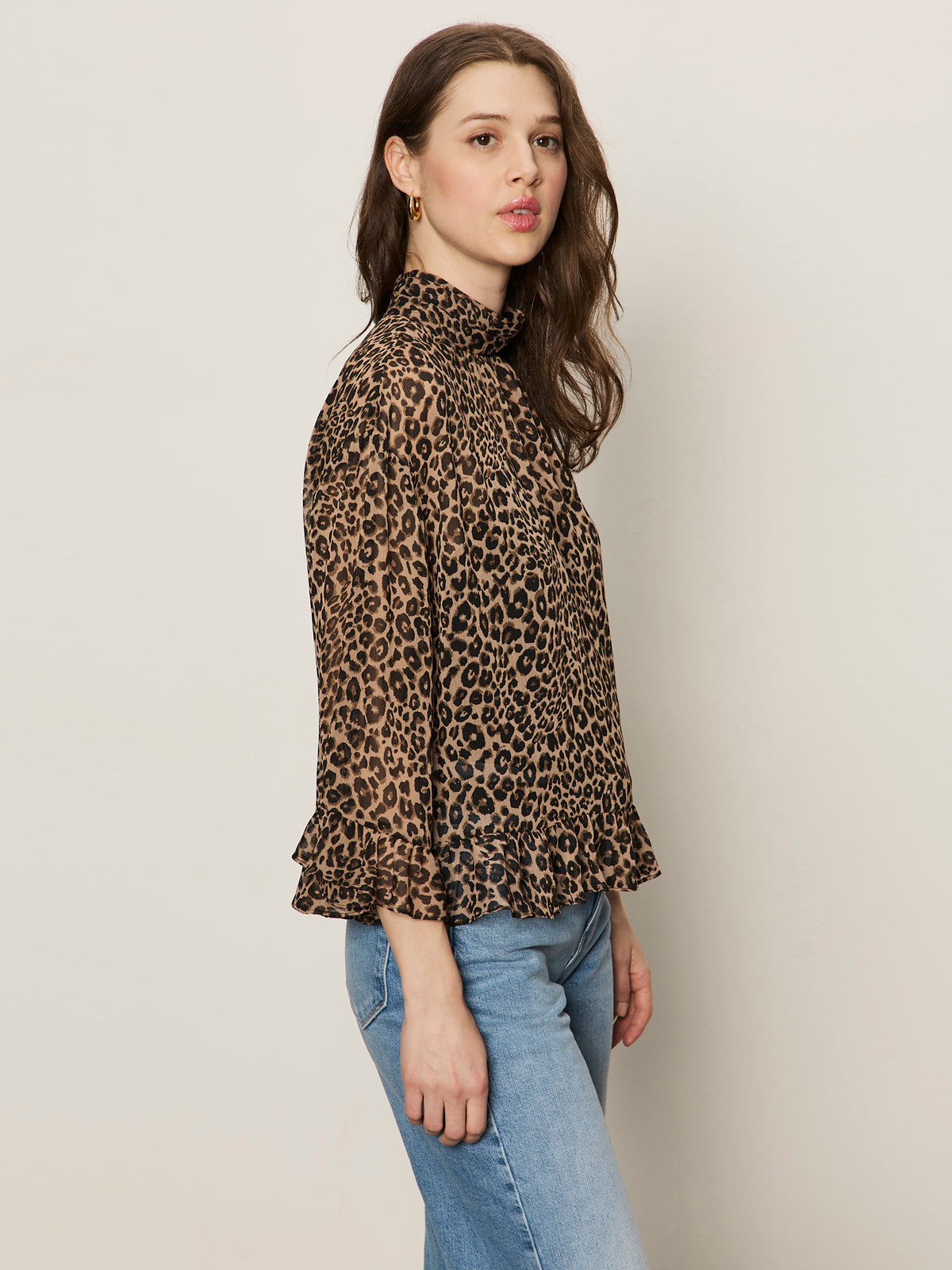 A woman with long brown hair wears Sanctuary Clothing's "the adore blouse french kitty," a high-neck, long-sleeved leopard print top with ruffled cuffs and hem, paired with light blue jeans against a plain light background.