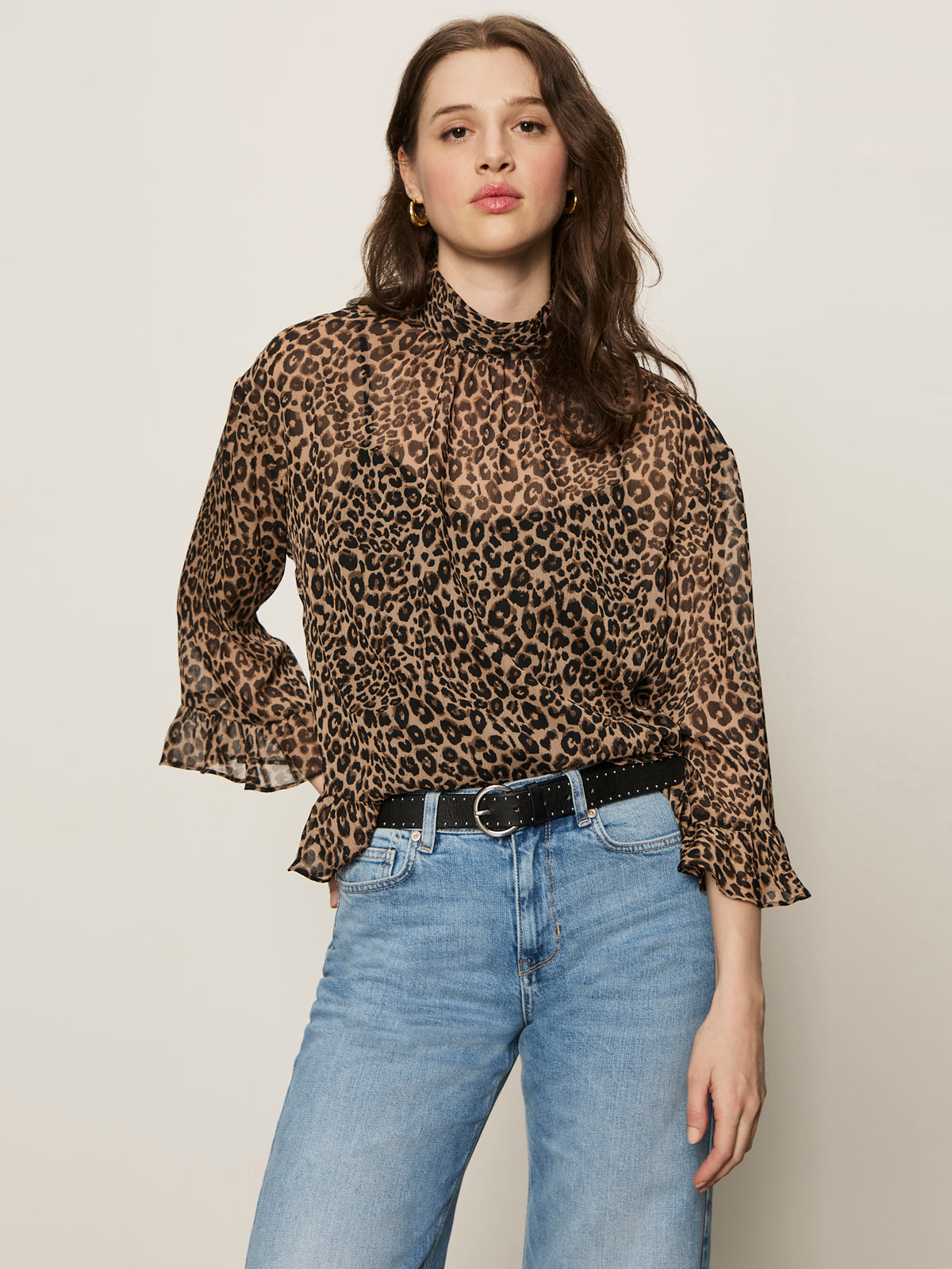 A woman with long brown hair models Sanctuary Clothing’s the adore blouse french kitty—a leopard print top with ruffled sleeves—paired with blue jeans, standing confidently against a plain light background.