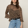 A woman with long brown hair models Sanctuary Clothing’s the adore blouse french kitty—a leopard print top with ruffled sleeves—paired with blue jeans, standing confidently against a plain light background.