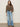 A woman poses against a plain background in the Adore Blouse French Kitty by Sanctuary Clothing— a loose, long-sleeved leopard print top—paired with high-waisted, wide-leg blue jeans and black sandals. One hand is in her pocket, hair loose and wavy.