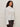 A woman with curly hair models the Sanctuary Clothing new era tuxedo shirt in white extended sizing, featuring pleated front details and wide cuffs, paired with dark pants against a plain light background.