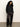 A woman with curly hair stands against a plain wall wearing the Sanctuary Clothing new era tuxedo shirt in black, paired with loose dark brown satin pants featuring a white side stripe. She looks neutral and relaxed.
