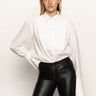 A woman with straight blonde hair models the Sanctuary Clothing new era tuxedo shirt in white, featuring wide cuffs, paired with black leather pants and one hand in her pocket against a plain white background.