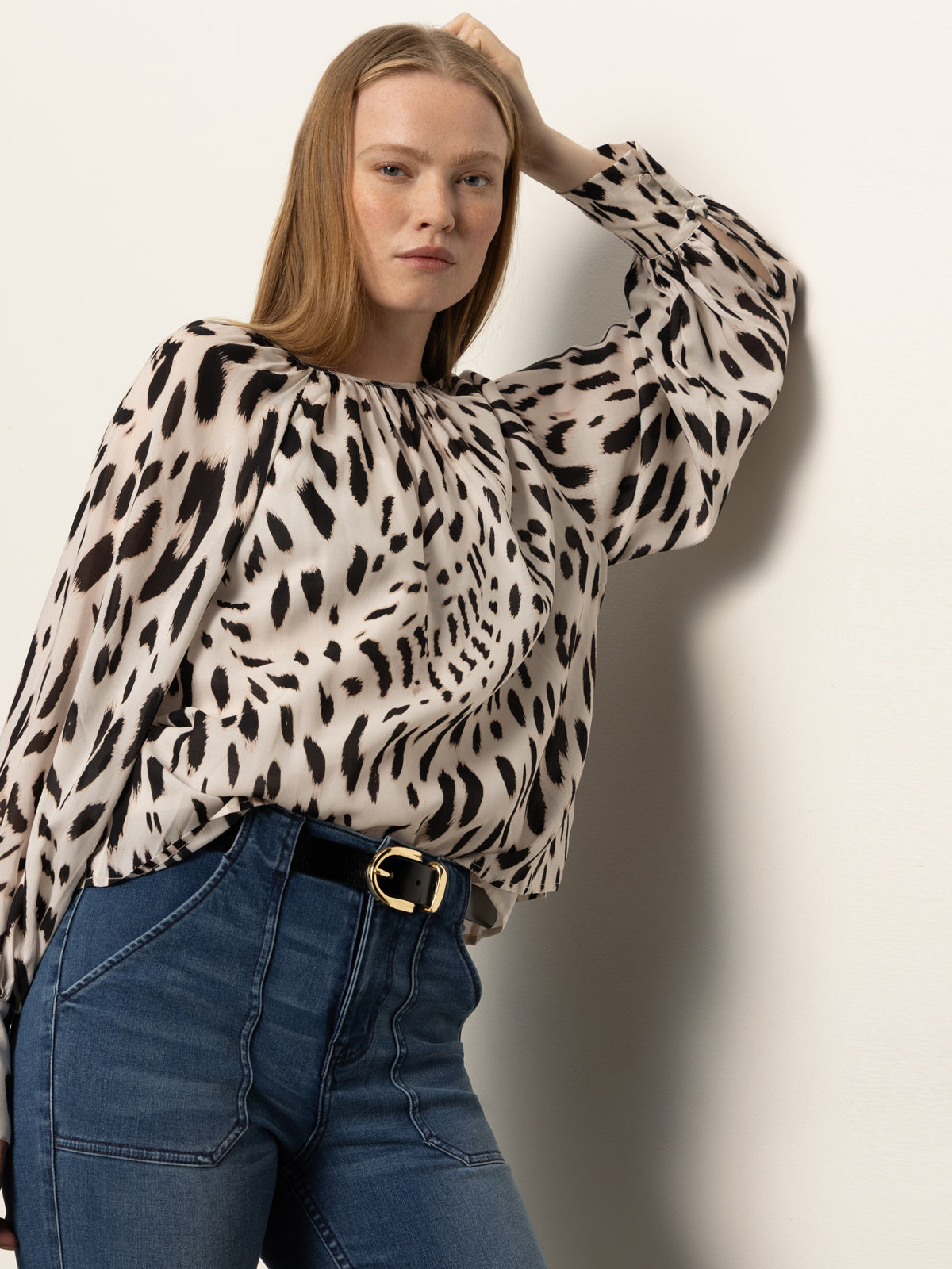 A woman with straight, light brown hair wears the Sanctuary Clothing Wild Dreamer Blouse Mother Leo—an animal print, long-sleeved top—tucked into high-waisted blue jeans with a black belt. She poses with one arm resting on her head against a light wall.