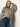 A woman with straight, light brown hair wears the Sanctuary Clothing Wild Dreamer Blouse Mother Leo—an animal print, long-sleeved top—tucked into high-waisted blue jeans with a black belt. She poses with one arm resting on her head against a light wall.