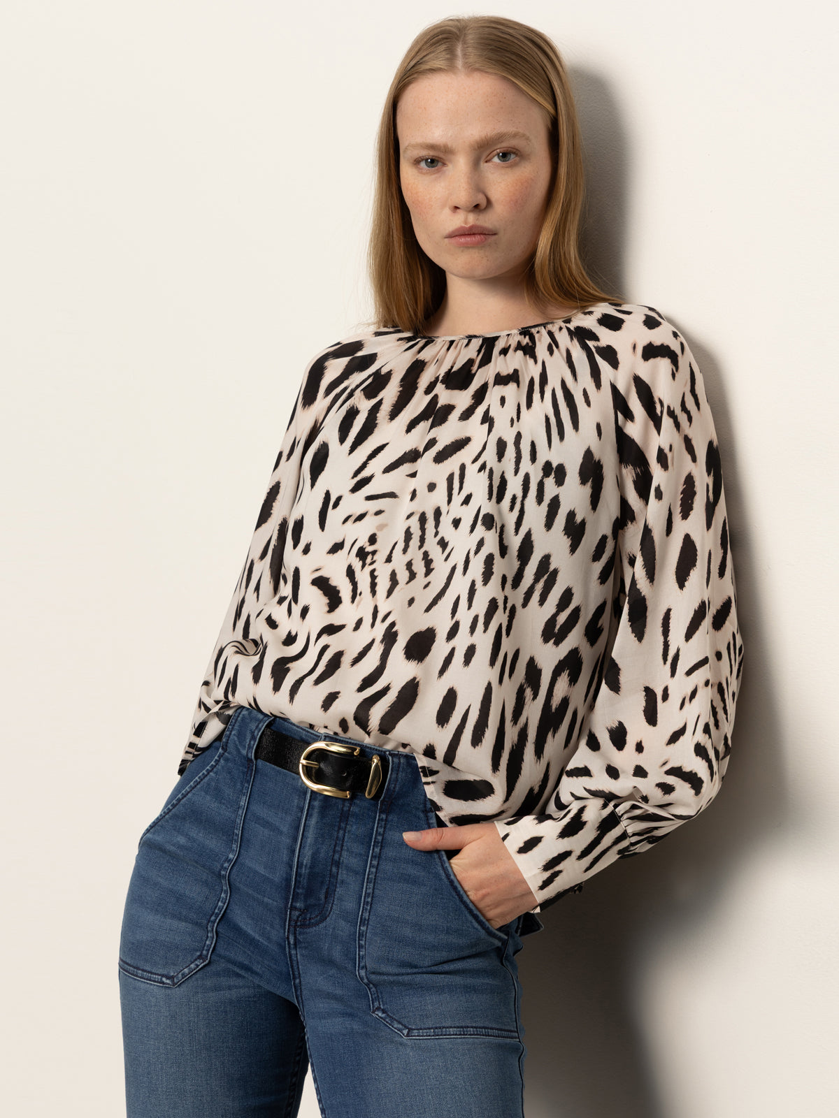 A woman with long, straight blonde hair wears Sanctuary Clothing's "wild dreamer blouse mother leo," a loose animal print top, tucked into high-waisted blue jeans with a black belt, standing against a plain light background and looking at the camera.