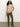 A woman with curly hair wears the Sanctuary Clothing sweet romance lace top fawn, styled with olive green pants and tan heeled boots, standing against a plain light background with one hand in her pocket.