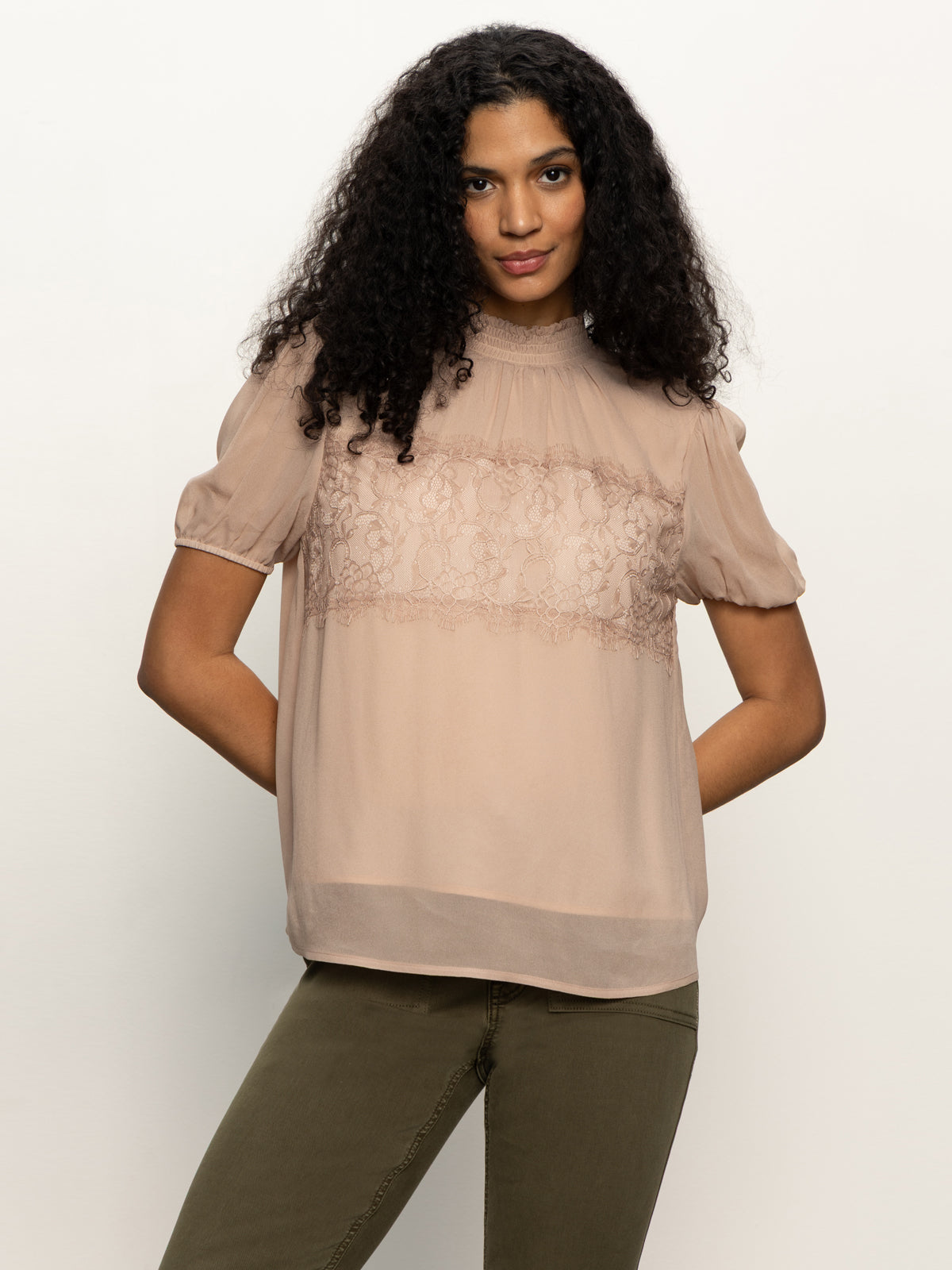 A woman with long curly hair smiles softly, wearing the Sanctuary Clothing sweet romance lace top in fawn and olive green pants, standing against a plain white background with her hands behind her back.