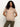 A woman with long curly hair smiles softly, wearing the Sanctuary Clothing sweet romance lace top in fawn and olive green pants, standing against a plain white background with her hands behind her back.