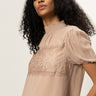 A woman with curly black hair models the Sanctuary Clothing sweet romance lace top in fawn, featuring short sleeves, lace chest detailing, and a high ruffled collar against a plain light background.