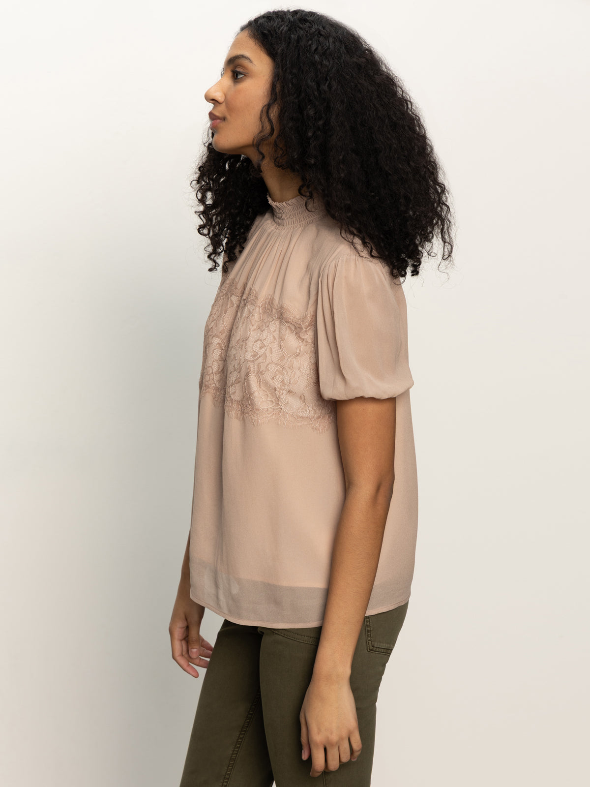 A woman with curly hair wears the Sanctuary Clothing sweet romance lace top in fawn, a light beige short-sleeve blouse with lace details, and olive green pants. She stands in side profile against a plain white background.