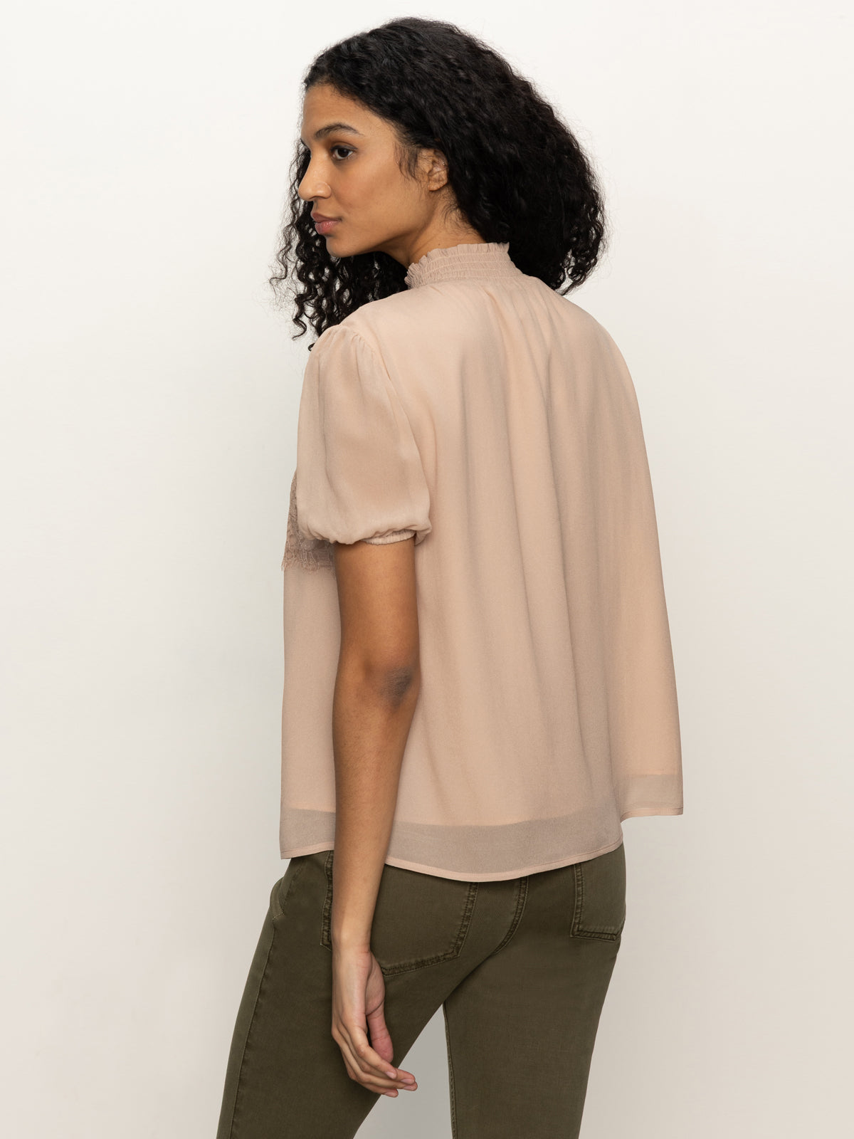 A woman with curly dark hair wears the Sanctuary Clothing Sweet Romance Lace Top in fawn, paired with olive green pants, standing against a plain light background and looking over her shoulder.