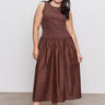 A woman with long dark hair wears Sanctuary Clothing's "take me away midi dress mesa" in extended sizing, featuring a sleeveless fitted top and gathered skirt, paired with black platform sandals. She stands on concrete before a plain light gray backdrop.