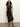 A woman with curly hair wears the Sanctuary Clothing everyday satin dress in black olive, standing against a plain white wall on a gray floor.