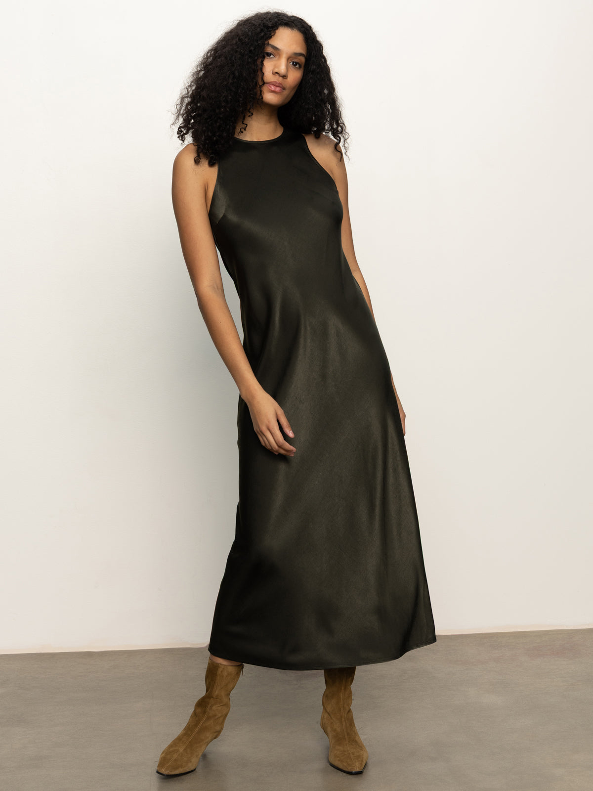 A woman with curly hair poses against a light wall, wearing the Sanctuary Clothing everyday satin dress in black olive and tan suede boots, with one hand on her hip.
