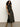 A woman with curly hair poses against a light wall, wearing the Sanctuary Clothing everyday satin dress in black olive and tan suede boots, with one hand on her hip.