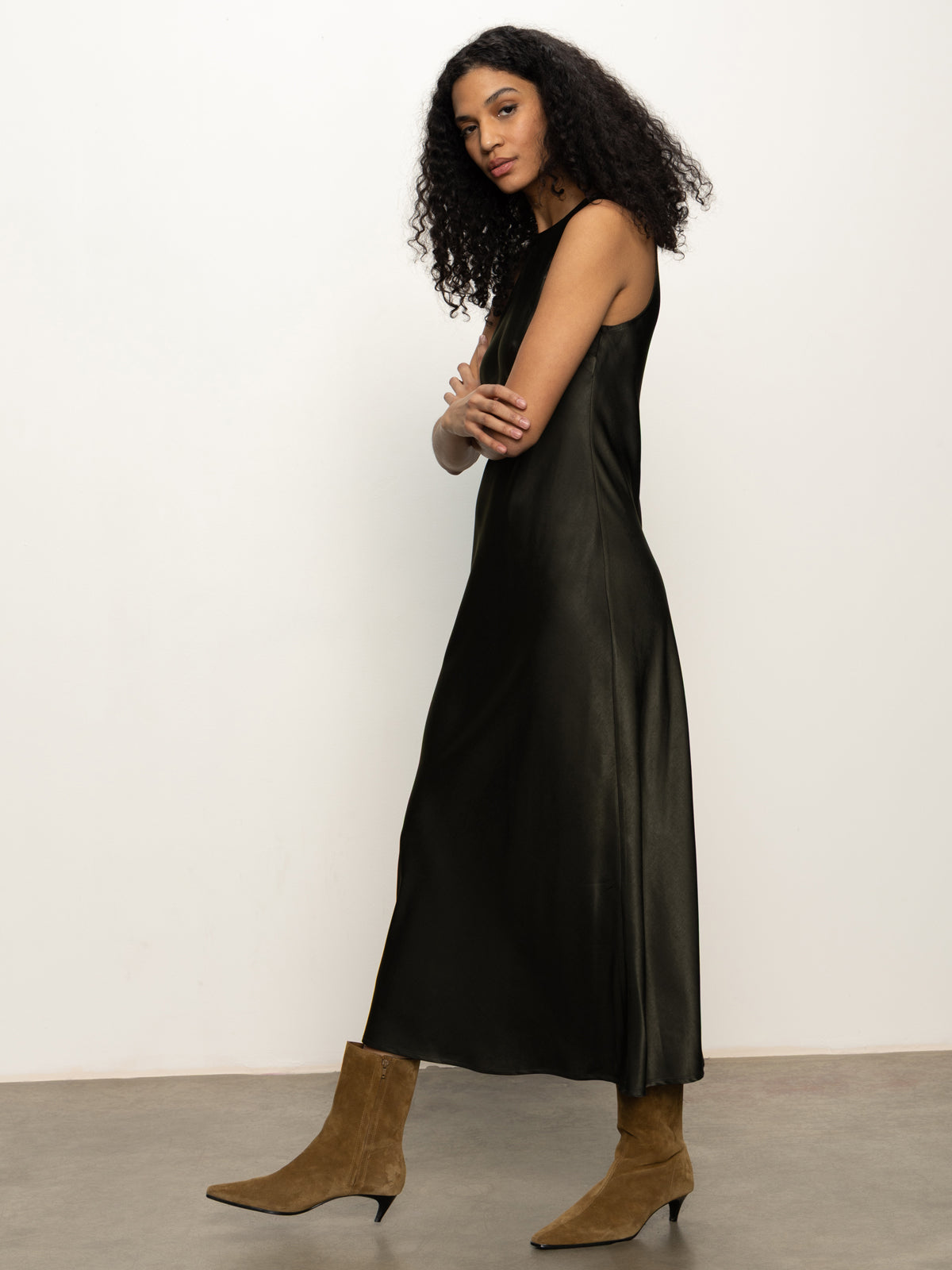 A woman with curly dark hair models the Sanctuary Clothing Everyday Satin Dress in black olive, paired with tan suede ankle boots. She stands sideways, arms crossed, looking at the camera against a plain white wall.