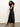 A woman with curly dark hair models the Sanctuary Clothing Everyday Satin Dress in black olive, paired with tan suede ankle boots. She stands sideways, arms crossed, looking at the camera against a plain white wall.