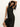 A woman with curly hair poses gracefully against a white background in the Sanctuary Clothing everyday satin dress in black olive, one arm raised and the other holding the dress near her knee.
