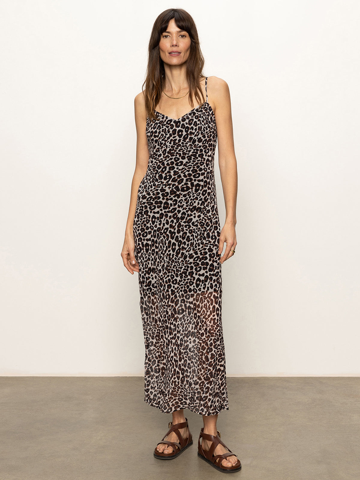 A woman with straight brown hair and bangs smiles slightly against a plain background, wearing the Sanctuary Clothing effortless slip dress meow—an ankle-length, sleeveless leopard print dress—with brown sandals.