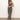A woman poses against a white wall, wearing Sanctuary Clothing’s effortless slip dress meow—an ankle-length, sleeveless leopard print dress—with brown platform sandals. She has long brown hair and rests one hand on her other wrist.