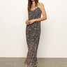 A woman poses against a white wall, wearing Sanctuary Clothing’s effortless slip dress meow—an ankle-length, sleeveless leopard print dress—with brown platform sandals. She has long brown hair and rests one hand on her other wrist.