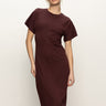 A woman with blonde hair models the Sanctuary Clothing cinched waist t shirt dress in chicory, featuring short sleeves and a midi length, standing against a plain light background with one hand on her hip.