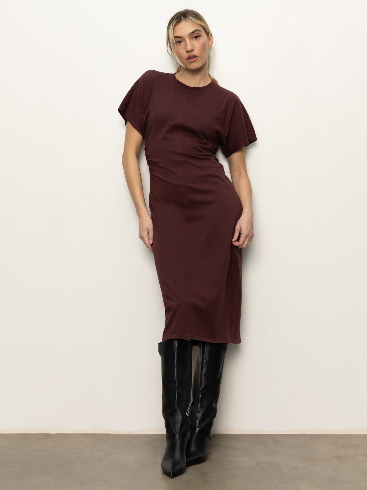 A woman with blonde hair pulled back poses against a light wall, wearing the Sanctuary Clothing cinched waist t shirt dress in chicory with tall black boots and one hand on her hip.