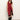 A woman in the Sanctuary Clothing revenge midi satin dress in crimson, featuring a sleeveless design and black lace-trimmed thigh-high slit, paired with knee-high black boots, stands against a plain light background.