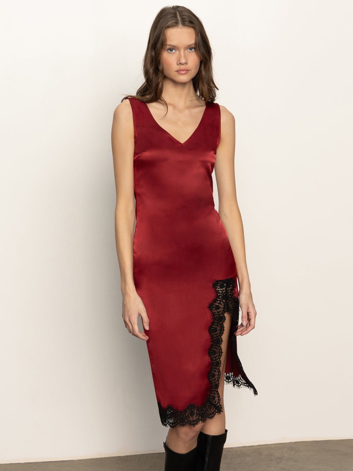 A woman wears the Sanctuary Clothing "revenge midi satin dress" in crimson, featuring a sleeveless V-neck, black lace-trimmed hem, and thigh-high slit, as she stands against a plain white wall.