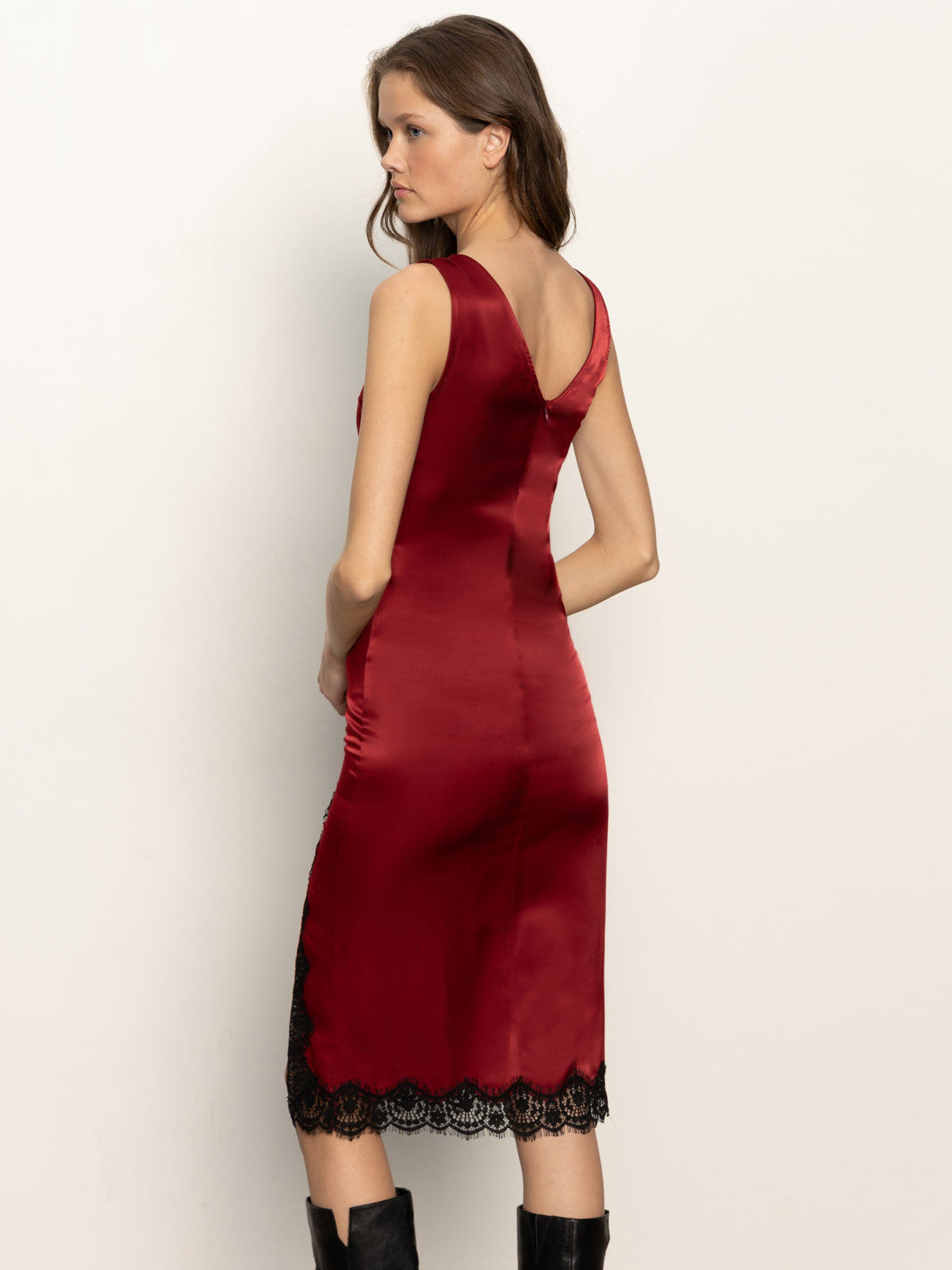 A woman stands sideways and glances over her shoulder, modeling the Sanctuary Clothing revenge midi satin dress in crimson. The sleeveless dress features black lace trim at the hem. She pairs it with knee-high black boots against a plain light background.