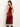 A woman stands sideways and glances over her shoulder, modeling the Sanctuary Clothing revenge midi satin dress in crimson. The sleeveless dress features black lace trim at the hem. She pairs it with knee-high black boots against a plain light background.