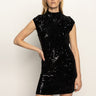 A woman poses against a plain light background in the Sanctuary Clothing "an affair to remember dress black," a sleeveless, high-neck sequin mini dress, with one hand on her hip.