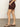 A person stands sideways against a plain light background and concrete floor, wearing Sanctuary Clothing's renegade short mesa—high-waisted brown shorts with rolled hems—and brown sandals with gold accents.