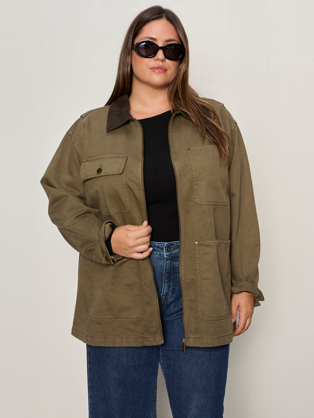 A woman with long brown hair wears Sanctuary Clothing's barn jacket in burnt olive (extended sizing) over a black top and blue jeans, paired with dark sunglasses, standing against a plain light background and looking at the camera.