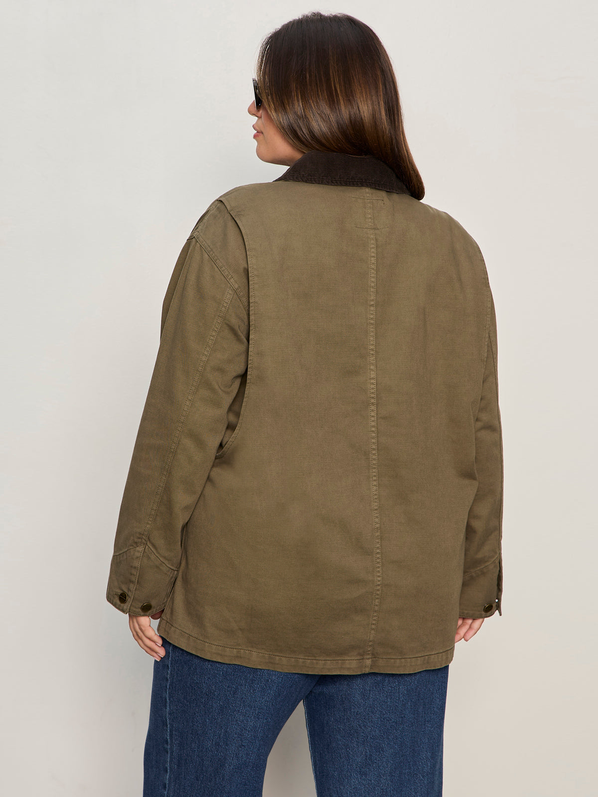 A woman with brown hair wearing sunglasses, blue jeans, and the Sanctuary Clothing barn jacket in burnt olive extended sizing stands with her back to the camera against a plain light background.