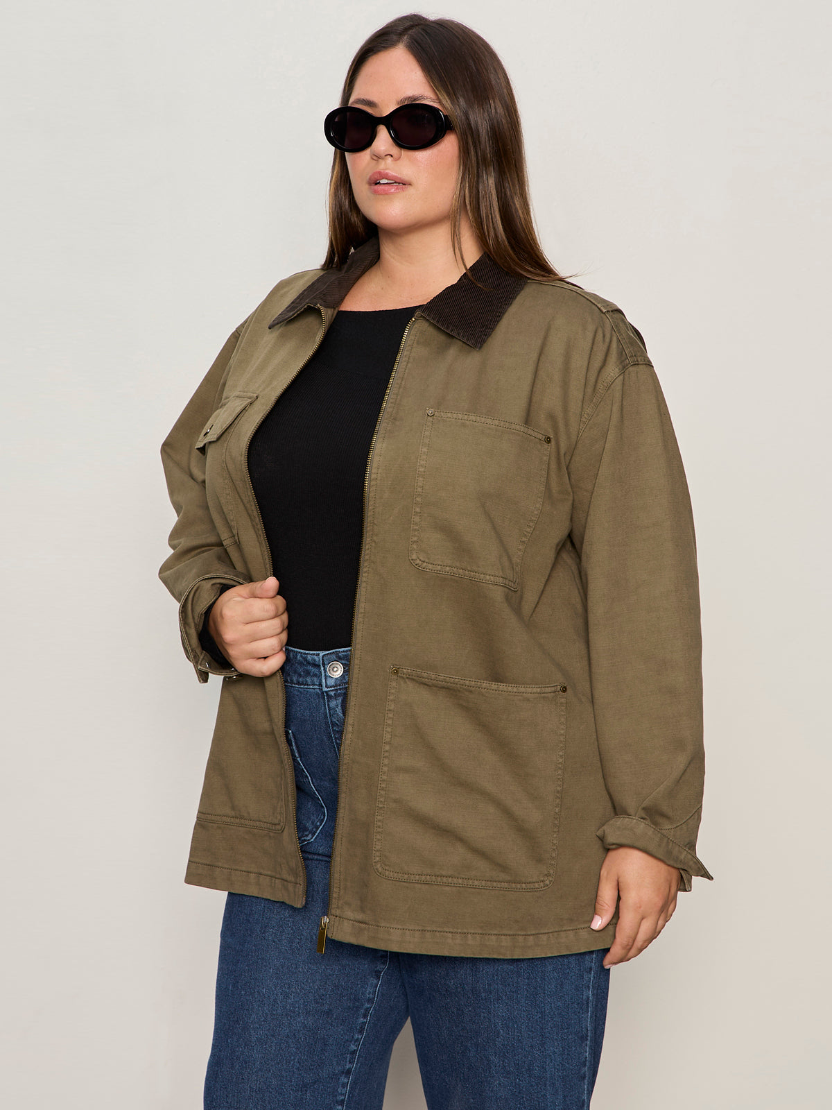 A woman in dark sunglasses, a black top, blue jeans, and the Sanctuary Clothing barn jacket burnt olive extended sizing stands against a plain light background.