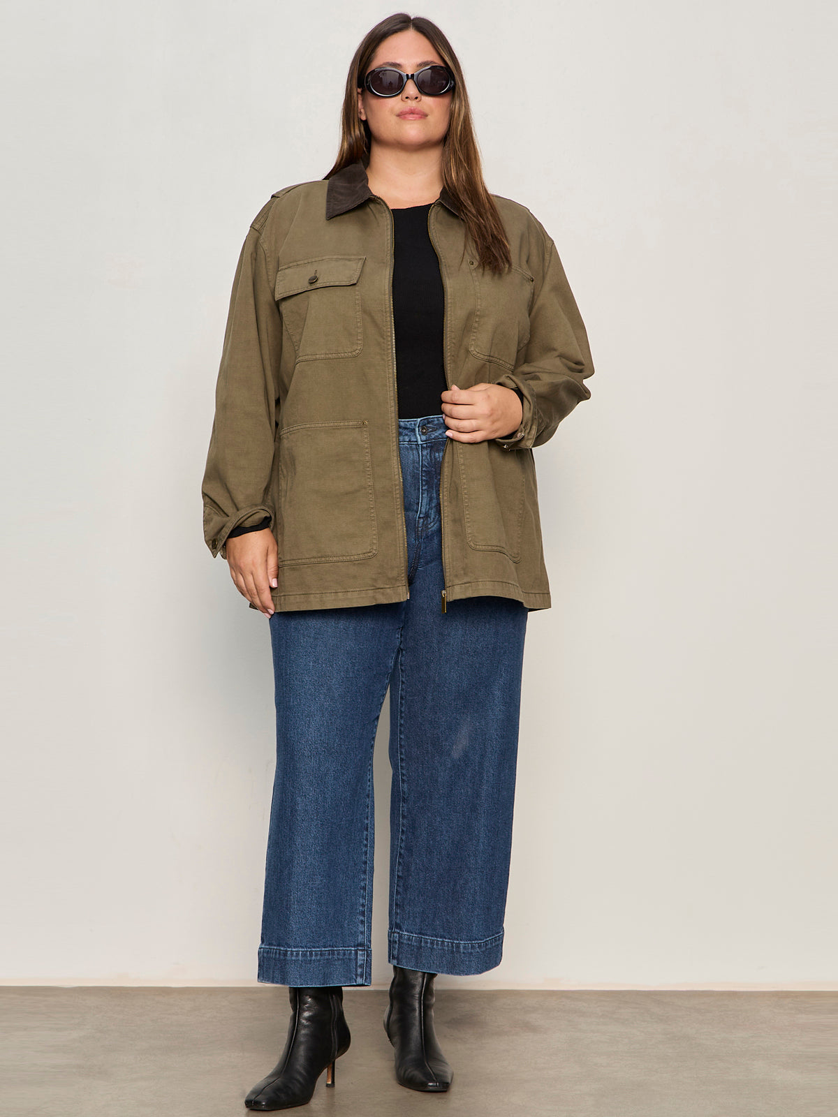A woman wearing sunglasses, a black top, wide-leg blue jeans, black ankle boots, and the Sanctuary Clothing barn jacket burnt olive extended sizing stands against a plain light-colored wall.