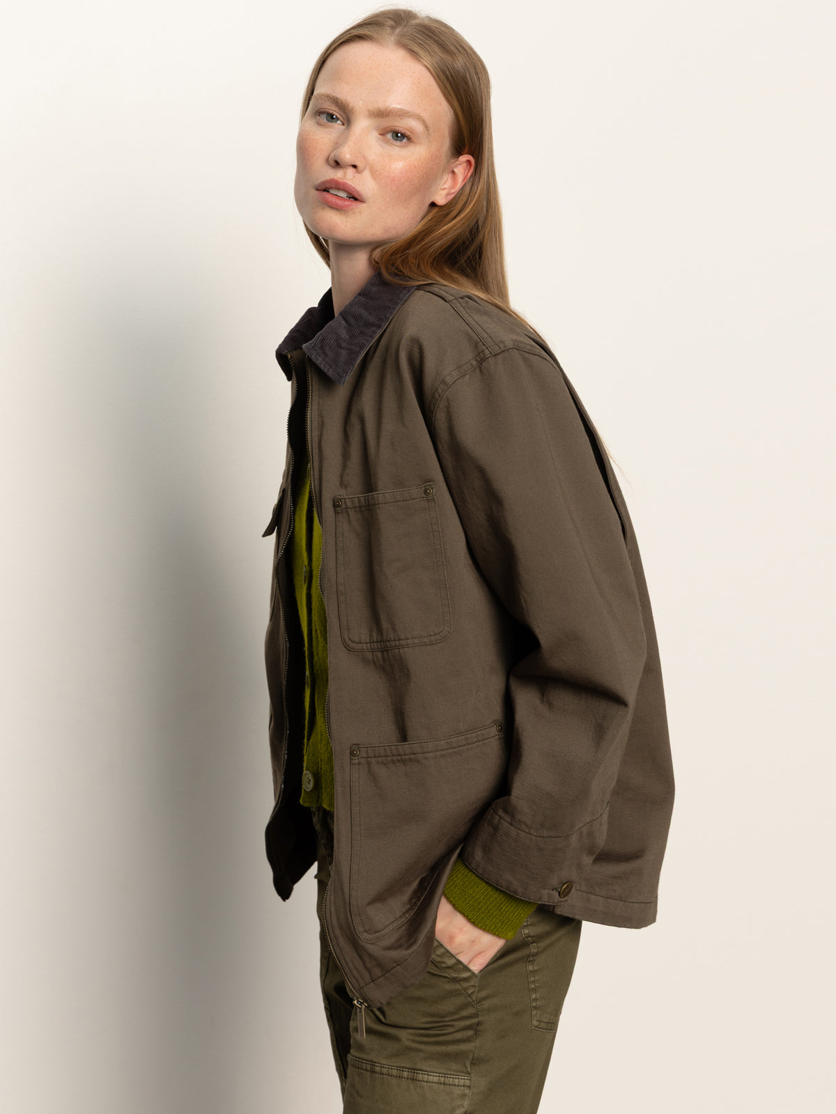 A person with long, straight, light brown hair wears the Sanctuary Clothing barn jacket in burnt olive over a green shirt and green pants, standing against a plain light background and looking slightly past the camera.