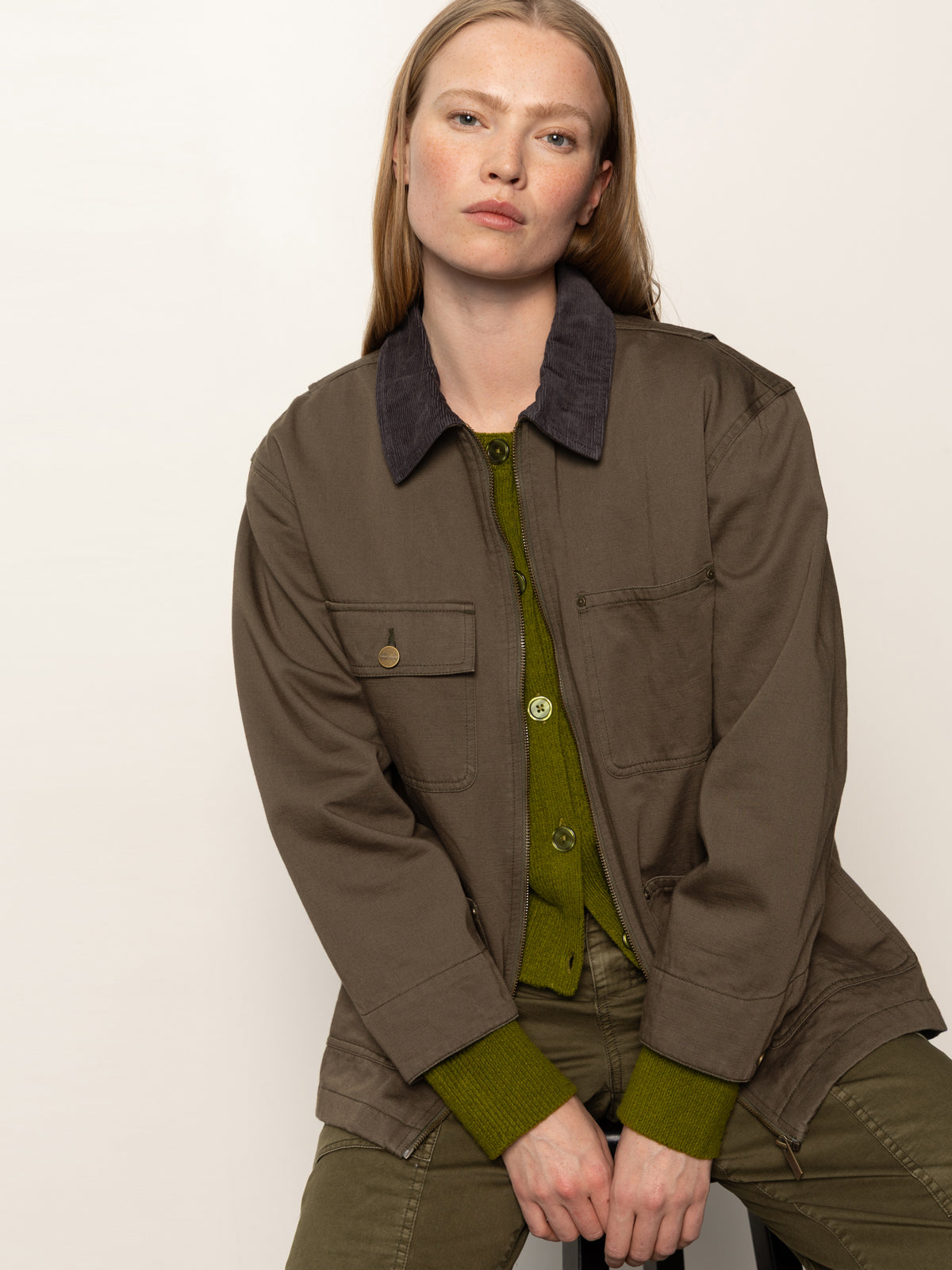 A woman with straight, light brown hair models the Sanctuary Clothing barn jacket in burnt olive over green separates, sitting against a plain background and looking directly at the camera with a neutral expression.