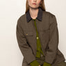 A woman with straight, light brown hair models the Sanctuary Clothing barn jacket in burnt olive over green separates, sitting against a plain background and looking directly at the camera with a neutral expression.