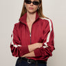 A woman in black sunglasses, dark jeans, and a Sanctuary Clothing satin track jacket in crimson with white sleeve stripes stands with arms crossed against a plain light background.